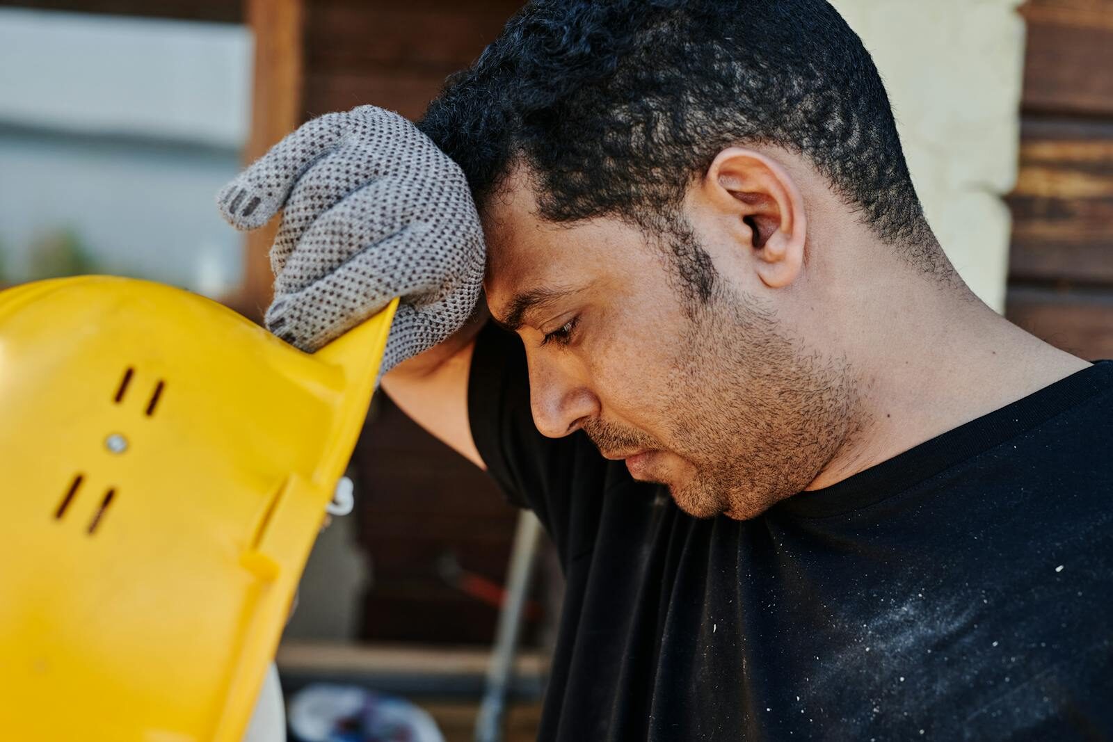 Side view of an adult male construction worker holding his hard hat, wearing gloves and deep in thought.